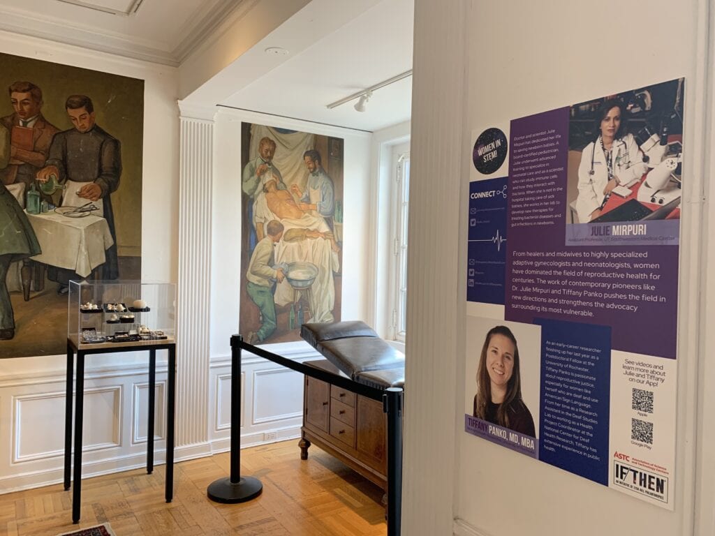 A museum exhibit featuring artwork, an old operating table, and a colorful wall display featuring women in science careers.